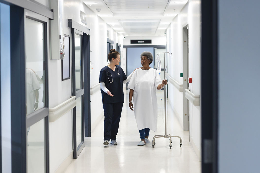 A nurse in navy-blue scrubs walks alongside an older patient wearing a white hospital gown in a bright hospital corridor. The patient holds an IV pole while the nurse gestures and speaks with them, suggesting support during a supervised walk or recovery exercise. The hallway is clean, well-lit, and lined with patient rooms on both sides.