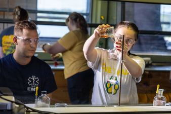 A student wearing safety glasses works inside a chemistry lab fume hood, carefully pouring liquid into a separatory funnel clamped to a stand. Laboratory glassware, beakers, and bottles are arranged on the work surface as the student focuses on the experiment. Sunlight from nearby windows illuminates the lab.