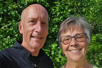 A smiling older couple stands outdoors in bright sunlight with lush green trees and shrubs behind them. The man, wearing a black quarter-zip pullover, and the woman, wearing purple-framed glasses and a light green top, are standing close together and looking at the camera. The photo is a close-up selfie with a vibrant, leafy background.